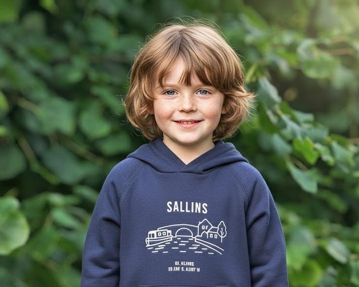 Child wearing a navy hoodie with 'Sallins' printed on it, standing in front of green foliage.